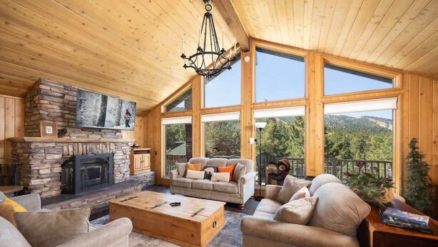A cozy living room with a stone fireplace, large windows, and a wooden ceiling.