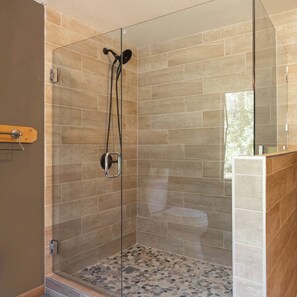 Ducha con paredes de vidrio, suelo con diseño de guijarros, banco de madera y espejo en la pared.