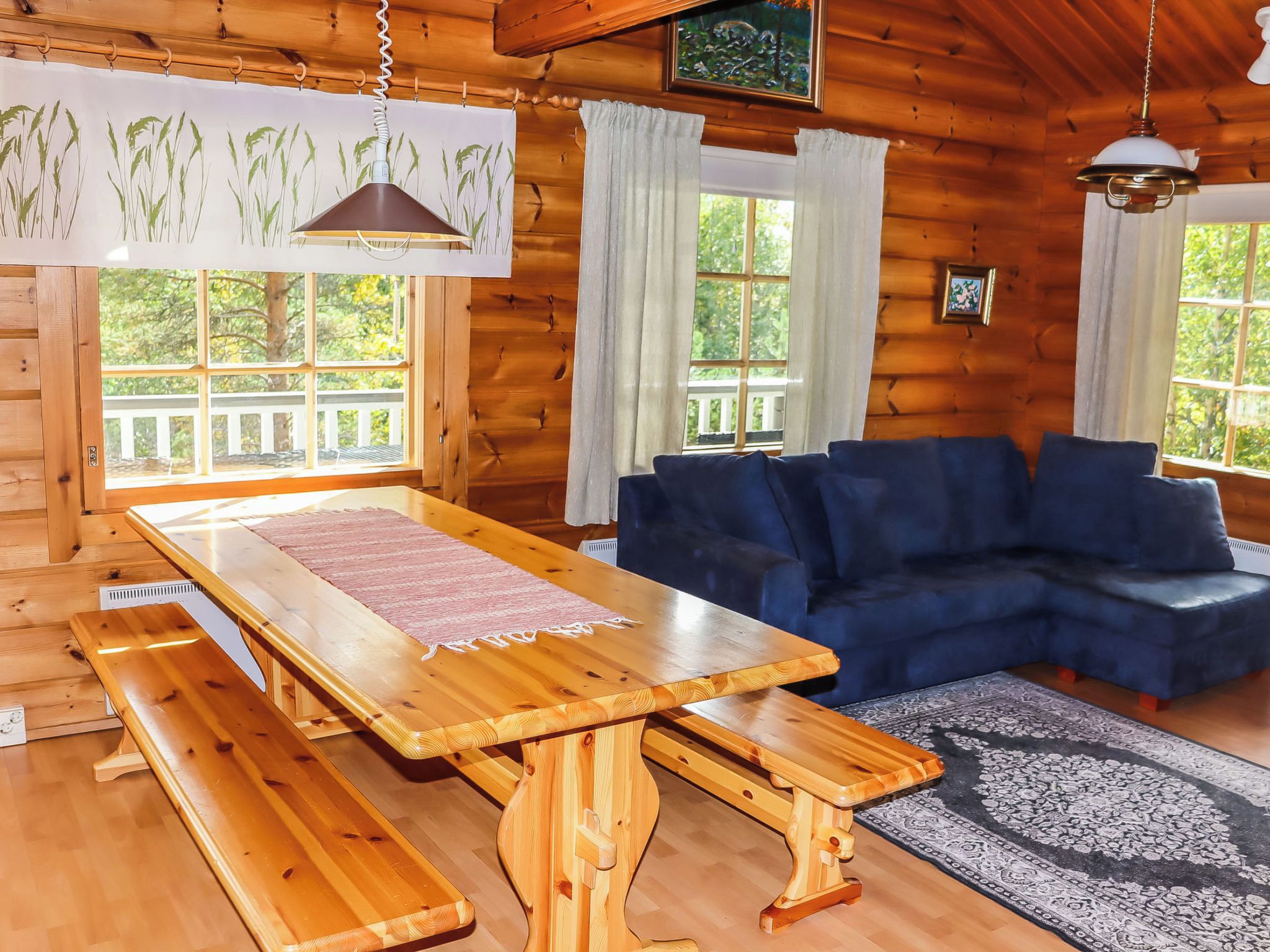 Interior de uma cabana de madeira com uma mesa de jantar, bancos, um sofá azul e janelas amplas.