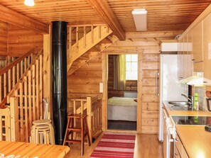 L’intérieur d’un chalet en bois comprend un escalier, un objet cylindrique noir, un lit, un coin cuisine et un tapis rouge.