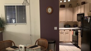 A dining area with a glass table and wicker chairs, a clock on the wall, and a kitchen in the background.