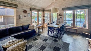 En rummelig opholdsstue med en stor sofa, et spisebord med stole og store vinduer med udsigt over en sø.