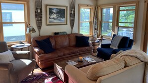 A cozy living room with a brown leather sofa, a beige armchair, and a coffee table. There are two windows with white frames and a painting on the wall. - LAKESIDE RETREAT!  A HIDDEN GEM!!! (Ossipee)