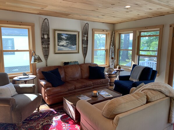 A cozy living room with a brown leather sofa, a beige armchair, and a coffee table. There are two windows with white frames and a painting on the wall. - LAKESIDE RETREAT!  A HIDDEN GEM!!! (Ossipee)