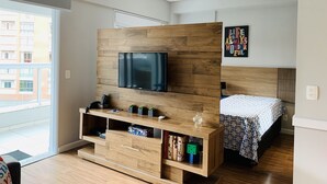 A modern bedroom with a wooden TV stand, a bed with patterned bedding, and a city view. - Akile Carlos Sampaio (São Paulo)