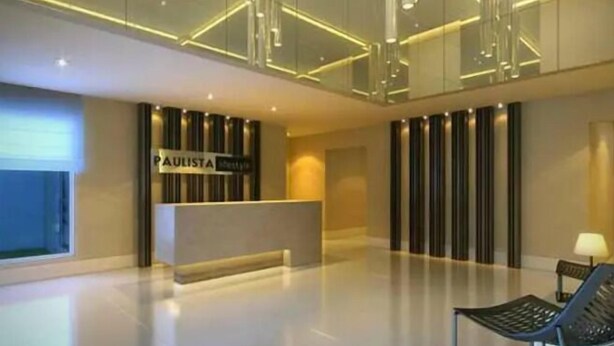 A modern reception area with a glass wall, a reception desk labeled "PAULISTA," and a seating area with a lamp. - Akile Carlos Sampaio (São Paulo)