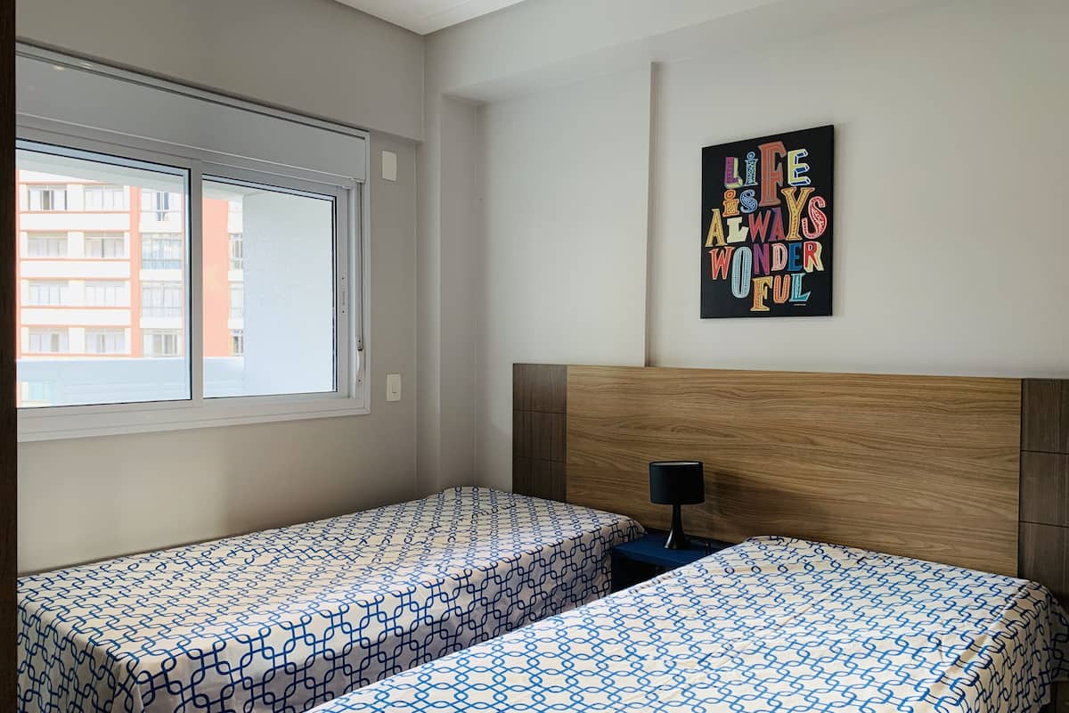 Habitación de hotel con dos camas individuales, cabecera de madera, ventana con persianas y un cartel motivacional en la pared.
