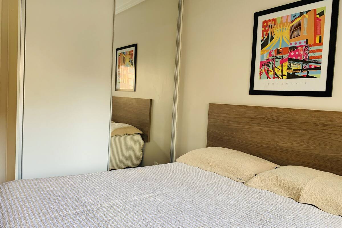 Un dormitorio con cabecera de madera, cama con cubierta blanca texturizada y un cuadro abstracto colorido en la pared.