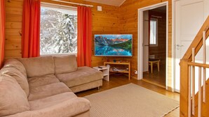 L’intérieur d’un chalet accueillant, avec un revêtement mural en bois, une grande fenêtre avec des rideaux, un téléviseur et un canapé.