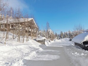Un sendero cubierto de nieve que conduce a un edificio con techo inclinado, rodeado de árboles cargados de nieve y un cielo azul despejado.