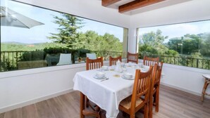 A dining area with a table set for six, large windows offering a view of greenery and trees, wooden beams on the ceiling, and a balcony with a glass railing. - Hercaris by Interhome (Begur)