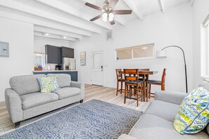 A modern living room with a grey sofa, a dining table with chairs, a ceiling fan, and a kitchen in the background.