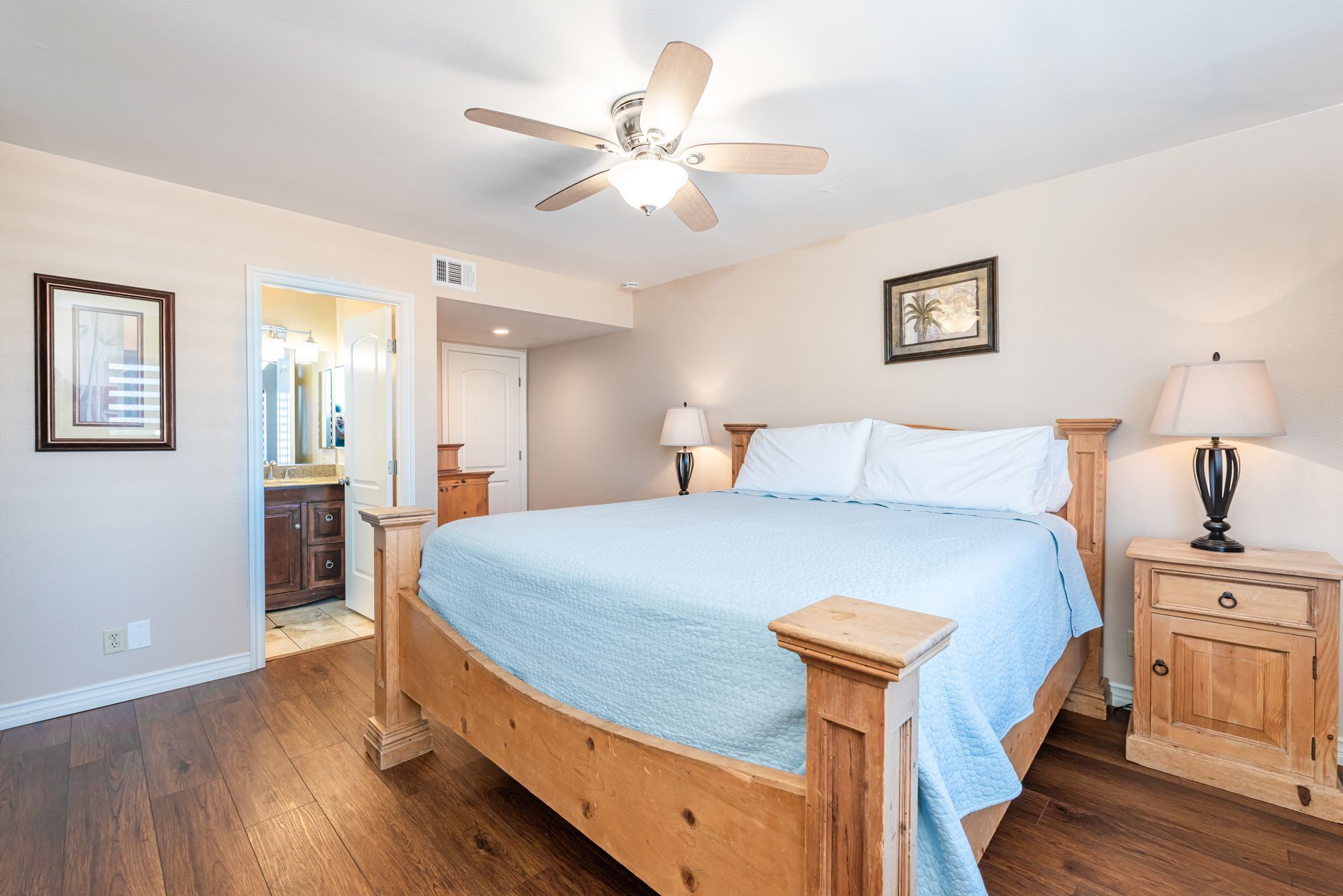 A bedroom with a wooden bed, a ceiling fan, a nightstand, a bedside table, and a door leading to a bathroom.