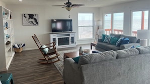A living room with a TV, rocking chair, and sofa. There is a bookshelf and a window with a view of the ocean. - AWESOME VIEW... On Private Gulf of Mexico Beach... Majestic Views  🐢🌴🌞 (Alligator Point)