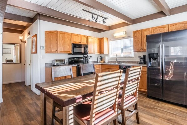 A modern kitchen with wooden cabinets, a stainless steel refrigerator, and a dining area with a wooden table and striped chairs.