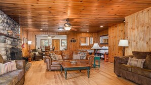 A cozy cabin interior with a stone fireplace, leather sofas, wooden floors, and a dining area.