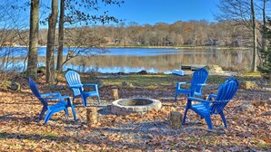 A lakeside seating area with blue Adirondack chairs around a stone fire pit, surrounded by trees and a clear view of the lake. - Lake Escape In the Poconos (Pocono Lake)