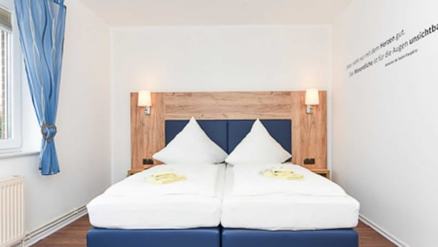 A double bed with a wooden headboard, two bedside lamps, and a window with blue curtains. - Double room C - Huus an't Deep in Altharlingersiel (Neuharlingersiel)