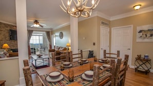 A dining area with a wooden table set for four, surrounded by rustic chairs. A chandelier hangs above, and a bear statue is visible in the background.