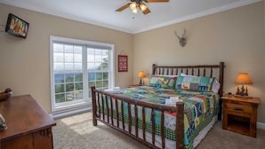 A bedroom with a large window, a ceiling fan, a bed with a colorful quilt, and a wooden nightstand.