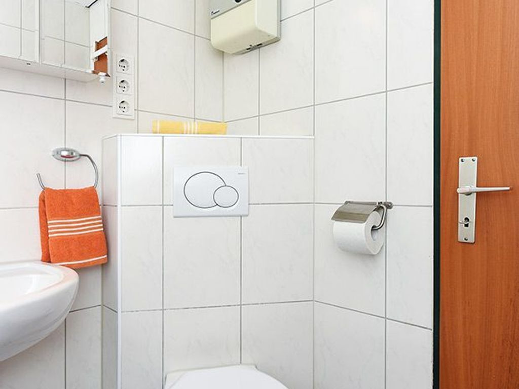 Une salle de bain avec un lavabo blanc, un porte-serviettes avec une serviette orange, un support à papier toilette et une porte dotée d’une poignée en métal.