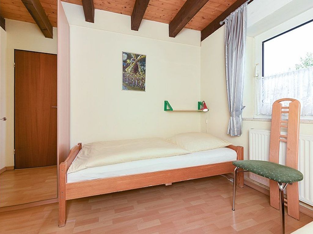 A single bed with a wooden frame, a small green shelf with a lamp, a chair, and a window with curtains.
