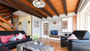 Ein Wohnzimmer mit einer Couch, einem Couchtisch und einem Fernseher. Im Hintergrund ist eine Treppe zu sehen.
