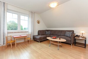 Uma sala de estar com um sofá marrom, duas cadeiras de madeira, uma mesa pequena e um abajur em uma mesinha lateral.