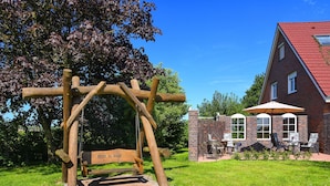Eine Holz-Pergola mit einer Bank, ein Backsteinhaus mit rotem Dach und Außenbestuhlung.