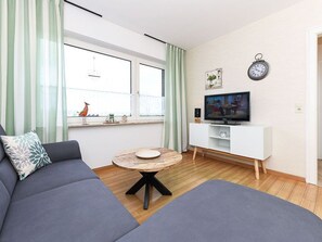A modern living room with a sofa, a round wooden coffee table, a television on a white cabinet, and a window with curtains.