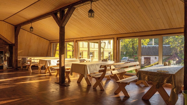 A spacious dining area with wooden benches and tables, large windows, and a high ceiling.