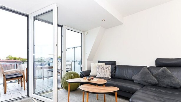 Een moderne woonkamer met een zwarte leren bank, twee houten bijzettafels en een groene poef. Een schuifdeur van glas leidt naar een balkon met uitzicht op de stad.