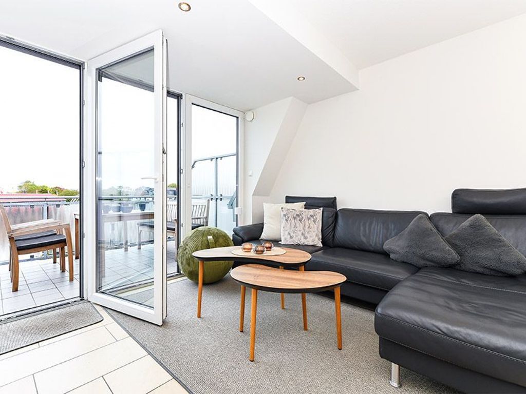 Un salon moderne avec un canapé en cuir noir, deux tables d’appoint en bois et un pouf vert. Une porte-fenêtre coulissante donne accès à un balcon extérieur offrant une vue sur la ville.