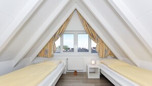 Une petite chambre mansardée triangulaire, avec deux lits simples, une table de chevet et un radiateur.