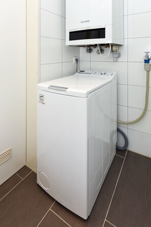 A compact, white front-loading washing machine in a small laundry room with white tiled walls and a brown floor.