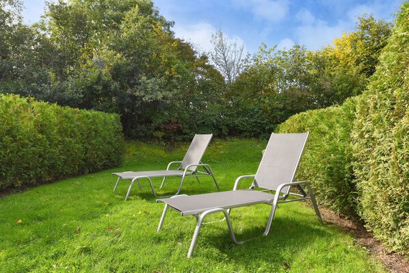 Two metal chairs with metal frames and grey fabric seats, placed on a grassy lawn with dense green hedges and trees in the background.