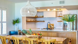 A modern kitchen with a wooden dining table, chairs, a fruit bowl, and a hanging light fixture. - Enjoy Modern Luxury Design in this Oceanfront, Corner Unit!  Stunning Ocean Views & Community Pool. (Folly Beach)