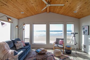 Una habitación acogedora con techo de madera, un ventanal grande con vistas a la playa, un sofá cómodo, una silla, un telescopio y una alfombra.