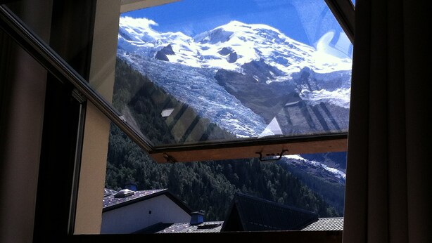 A view through a window of a mountain landscape with snow-covered peaks and a forested valley below.