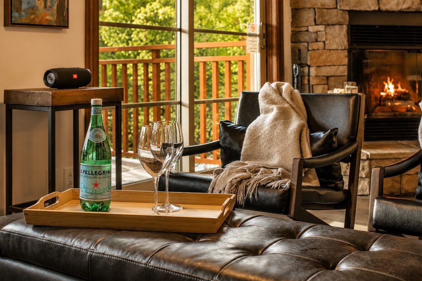 Un salon chaleureux avec un canapé en cuir, une table basse en bois sur laquelle se trouvent une bouteille et des verres, une cheminée et une vue sur la verdure à l’extérieur.