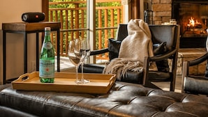 Un salon chaleureux avec un canapé en cuir, une table basse en bois sur laquelle se trouvent une bouteille et des verres, une cheminée et une vue sur la verdure à l’extérieur.