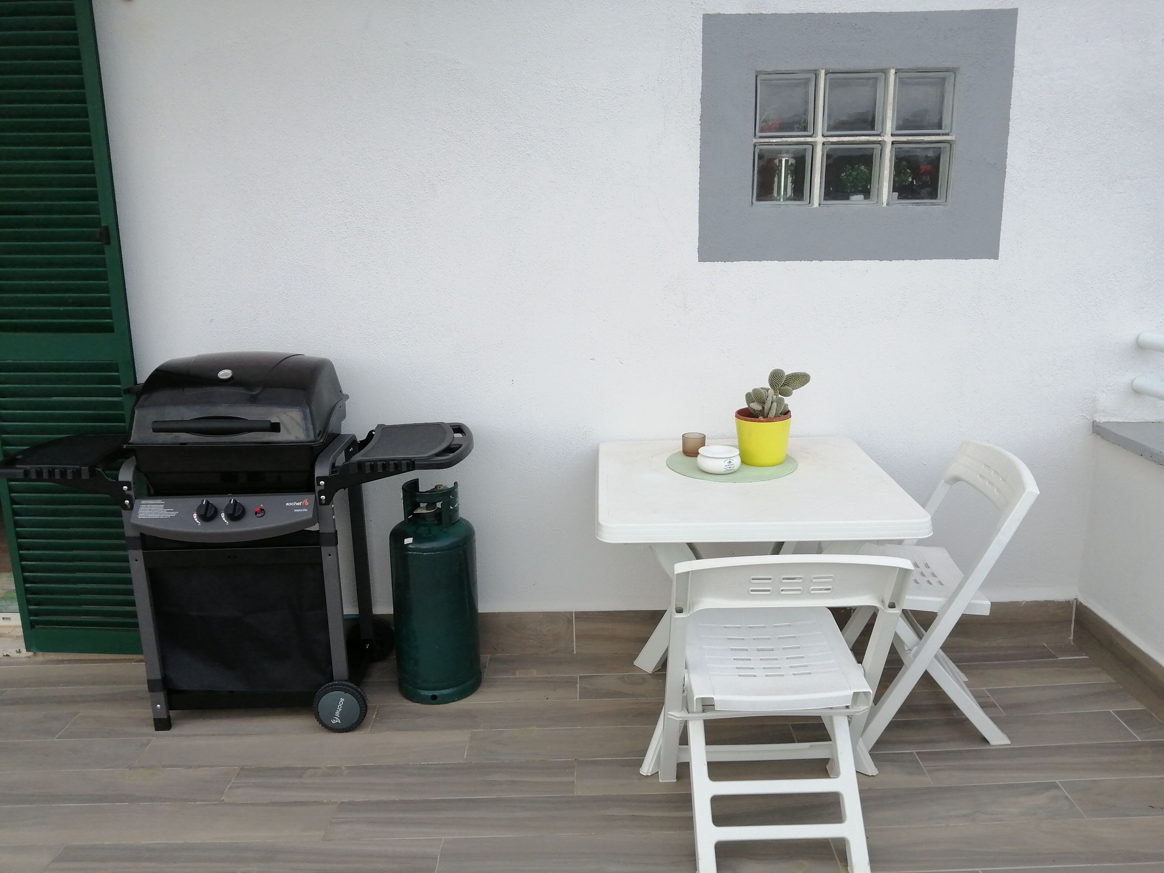 A small outdoor seating area with a white table, a single white chair, a small potted plant, a grill, and a gas cylinder.