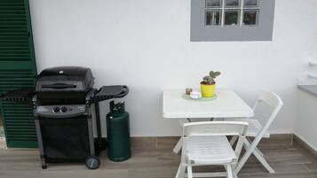 A small outdoor seating area with a white table, a single white chair, a small potted plant, a grill, and a gas cylinder.