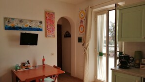Ein Essbereich mit Holztisch und Stühlen, einem an der Wand befestigten Fernseher und einer Küchenzeile mit einer Kaffeemaschine.