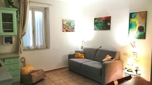 Ein Wohnzimmer mit einem grauen Sofa, einem kleinen runden Tisch mit einer Lampe und einem Fenster mit Vorhängen.