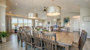 A spacious dining area with a large wooden table and matching chairs, overhead lighting, and a view of the outdoors through large windows.