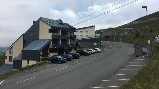 A parking lot with several cars, a building with a sloped roof, and a hillside in the background.
