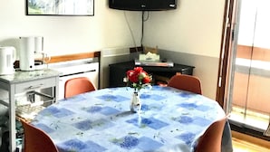 A dining area with a table covered in a floral tablecloth, surrounded by orange chairs. A vase with red flowers is on the table. A flat-screen TV is mounted on the wall above the table.