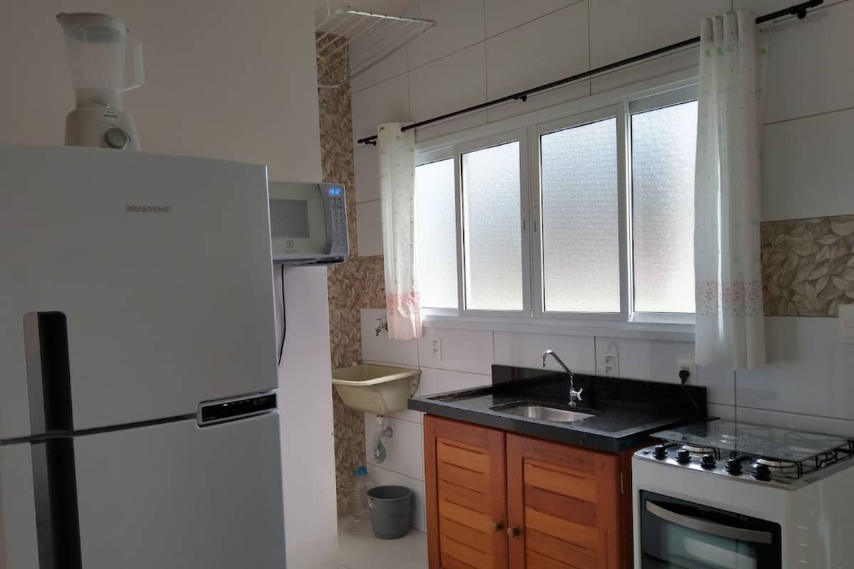 A kitchen with a refrigerator, microwave, sink, wooden cabinets, and a window with curtains.