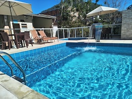 Uma piscina com água azul cristalina, cercada por cadeiras de espreguiçadeira e guarda-sóis, com vista para uma montanha e árvores ao fundo.